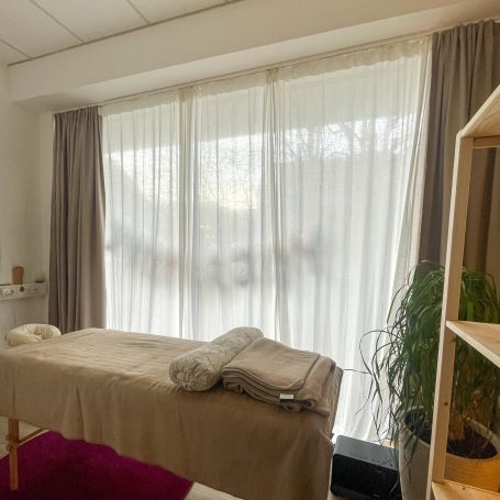 Heller Wellnessraum mit einem Behandlungstisch und Fenster mit Vorhängen.